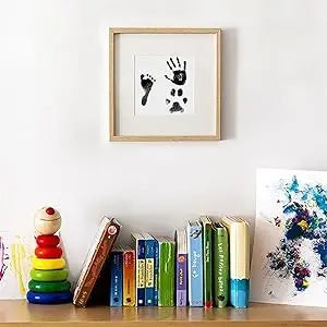 image of a photo frame with examples of black prints including a Childs foot a child hand and an animal paw print  located above a book shelf with a selection of colourful books underneath.